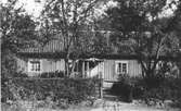 Bostadshus vid Onsalavägen från Kungsbacka. Fönstret till höger om den lilla farstukvisten står öppet. Trädgård hägnas med en låg stenmur och häck.