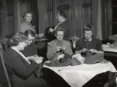 Beredskapsstickning på Nordiska kompaniet. Fyra kvinnor sitter runt bord och stickar, två kvinnor står bakom. 