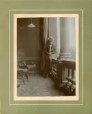 Bib Adelsköld fotograferad i hatt och tvådelad klänning. Fotografiet är taget på Villagatan 2 i Stockholm där familjen bodde. Bib sitter på ett räcke till en loggia/balkong ut mot Karlavägen. På balkongen syns två korgstolar samt ett mosaikgolv.