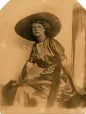 Bib Adelsköld fotograferad i sidenklänning och bredbrättad flätad hatt. Fotografiet är taget 1912 och har en otydlig signatur.