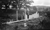 Två män arbetar med att slå ner en spåntvägg tvärs över den inre vallgraven vid Rumlaborg i Huskvarna sommaren 1932, i samband med arkeologiska undersökningar på platsen. Spåntväggens funktion var att hindra vatten från Huskvarnaån att rinna in i vallgraven.
