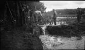 Några män och skolpojkar står vid en fördämning mellan Huskvarnaån och den inre vallgraven vid Rumlaborg i Huskvarna. Fotot togs i samband med arkeologiska undersökningar vid borgen sommaren 1935.
