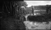 Några män och skolpojkar står vid en fördämning mellan Huskvarnaån och den inre vallgraven vid Rumlaborg i Huskvarna. Fotot togs i samband med arkeologiska undersökningar vid borgen sommaren 1935.
