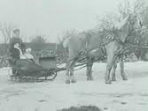 Ett par hästar och släde. Hästarna har bjällror.  I släden sitter en kvinna och ett barn. Bak på släden står en kusk som håller i en piska. I bakgrunden en stengärdsgård och bortom den syns taket på ett hus.