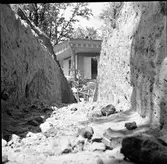 Två personer står i ändan av schakt C genom den södra delen av borgkullen vid Rumlaborg i Huskvarna. I bakgrunden syns lusthuset på toppen av kullen. Fotot är taget i samband med arkeologiska undersökningar vid borgen 1937. Vid fototillfället grävs det andra lagret, av totalt tre i schaktet, bort.