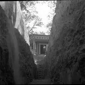 En man står ovanför schakt C som grävts genom den södra delen av borgkullen vid Rumlaborg i Huskvarna. I bakgrunden syns lusthuset som står på toppen av kullen. Vid fototillfället har den tredje, av totalt tre utschaktningar påbörjats. På den djupaste delen, närmast lusthuset kom schaktet att bli ca 4,5 meter djupt. Fotot är taget i samband med arkeologiska undersökningar vid borgen sommaren 1937.