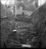 Schakt D genom den södra delen av borgkullen vid Rumlaborg i Huskvarna i samband med arkeologiska undersökningar sommaren 1938. Fotot är taget mot söder och i förgrunden kan stockrester ses, lite längre fram även rester av en pålrad. I bakgrunden står tre män, vid sidan av pumpen som användes för att hålla schaktet någorlunda torrt. Rester efter pålrader påträffades även vid andra delar längs med borgkullens kant i samband med de arkeologiska undersökningarna, som bedrevs av Huskvarna hembygdsförening mellan åren 1931-1942.