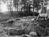 Grävning av schakt F på östra sidan av den inre vallgraven vid Rumlaborg i Huskvarna sommaren 1939.  Till höger om schaktet står en ung man bakom en sållställning. I bakgrunden syns borgkullen med lusthuset på toppen. Fotot är taget i samband med arkeologiska undersökningar av borgen, bedrivna av Huskvarna hembygdsförening.