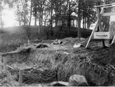Grävning av schakt F på östra sidan av den inre vallgraven vid Rumlaborg i Huskvarna sommaren 1939.  Till höger om schaktet står en ung man bakom en sållställning. I bakgrunden syns borgkullen med lusthuset på toppen. Fotot är taget i samband med arkeologiska undersökningar av borgen, bedrivna av Huskvarna hembygdsförening.