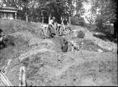 Fyra skolpojkar och en man från Huskvarna stad gräver i schakt G vid sydvästra sluttningen av borgkullen vid Rumlaborg i Huskvarna sommaren 1942. I bakgrunden syns lusthuset på kullens topp.