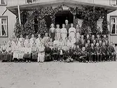 Fotografi från Kungsäter. En stor grupp ungdomar sittande vid en veranda; pojkar till höger och flickor till vänster. Uppe på verandan står en vuxen man, präst eller lärare förmodligen. Det kan vara en skolklass eller konfirmandgrupp. Verandan är inklädd med någon slingerväxt.