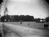 Varbergs torg sett från Kungsgatan mot kyrkan och till höger den så kallade brännvinsringen från 1880-talet. På dess stensatta sockel satt torgmadamerna och sålde sina varor. Till höger står tre små flickor och fler personer rör sig på torget. I hörnbyggnaden till höger om kyrkan låg det gamla apoteket.