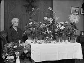 Kvinna i hemmiljö omgiven av blommor, Uppland 1948