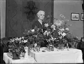 Kvinna i hemmiljö omgiven av blommor, Uppland 1948