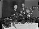 Kvinna  och man i hemmiljö omgiven av blommor, Uppland 1948