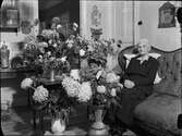 Kvinna i hemmiljö omgiven av blommor, Uppland 1948