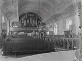 Interiörbild av Varbergs kyrka före renoveringen 1932, då bl a dekormålningarna målades över och bänkarna ändrades. Vy från koret mot orgelläktaren. 
(Skada i höger sida av fotografiet)
