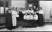Gruppbild av 2 män och 12 kvinnor anställda vid en syverkstad i Alingsås. Möjligen Nygatan 24. På dörren bakom gruppen står det Doktor A. Hedberg.