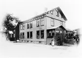 Gatuvy av Café Göta när det låg på Nygatan, kv. Färgaren 5, mellan 1889-1930. Caféet låg på första våningen, vid ingången står en kvinna i förkläde samt två män. På innegården syns människor samt en häst. En skylt på fasaden med text 
