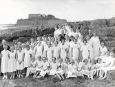 En sångkör på utflykt till badplatsen Djupa Dräkt. Folkskolans barnkör i vita kläder uppställda vid stranden med folkskolärare Österberg i mitten och Varbergs fästning i bakgrunden.