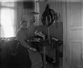 Gunnars mor Bertha vid stickmaskinen. Bertha har skyddat tapeten mot de skador stickningen kan åstadkomma och kvällstid har hon endast en fotogenlampa som belysning. Troligen ryms maskinen i köket eftersom det hänger brickor i brickband på väggen över den. 

I Viskadalen, och Sjuhäradsbygden i övrigt, förekom det s k förläggarsystemet. Förläggarna köpte levererade material och utrustning till textilarbeterskorna i hemmen runtom i trakten. Förläggarna samlade sedan in de färdiga produkterna.