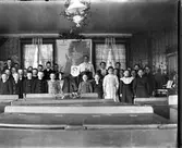 Lärarinnan Constance Engström (fotografens moster) med en klass samlade vid katedern i Sällstorps skola. I förgrunden skolbänkar och på katedern står ett fotografi av en man insvept i tyg och blomstersmyckning. Mellan fönstren hänger en stor karta över Palestina.