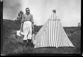 En kvinna står vid ett randigt tält på Maltaremosse. I ena handen håller hon en mjölkkanna och i den andra har hon en korg, troligen med mat till tältaren. På tälttoppen sitter en hatt. I bakgrunden till vänster ses murare Andreas väderkvarn.