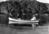 En finklädd familj på båttur med anordning för laxfisket vid Åsbro i bakgrunden och ett brant berg. I aktern sitter en kvinna i hatt med ett litet barn och styr båten. I fören sitter en man i skärmmössa och längst fram är en flagga monterats.
