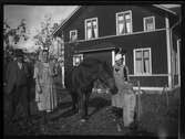 Grupporträtt, personer och häst, stående framför husgavel.
