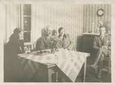 Fyra personer sitter vid ett bord i ett kök/rum, okänd plats och årtal. Foto från Sagered-album.