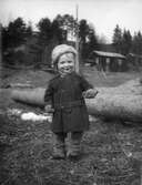 Ett barn framför några timmerstockar. I bakgrunden träd och byggnader. Resele cirka 1925.