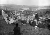 En man som möjligen kan vara en präst står och läser för en grupp människor som sitter i gräset. I bakgrunden en vy med Ångermanälven och bebyggelse. Resele omkring 1925.