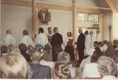 Vid invigningen av Apelgårdens kyrka (Svenska kyrkan) 1982. Biskop Bertil Gärtner står vid altartavlan. I folkdräkt står Margit Johansson, Torrekulla.