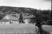 Grimmared, Låbäck, bostads- och uthus som alla är omålade. Bakom gården reser sig ett berg. Utanför stugans farstukvist står husbonden och makan sitter bredvid honom med ett litet barn i knät. Åkrarna går nära in på husen.