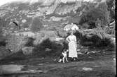 En kvinna står med en stor hund i naturen. I bakgrunden reser sig en hög bergvägg och ett hus ligger rakt bakom kvinnan.