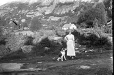 En kvinna står med en stor hund i naturen. I bakgrunden reser sig en hög bergvägg och ett hus ligger rakt bakom kvinnan.
