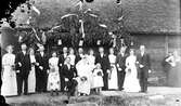 Svennerbergs bröllop, ca 1914 i Svenstorp, Larsa, Sällstorp. Brudparet sitter bredvid sina brudnäbbar och framför sina gäster, tärnor och marskalker. De är samlade vid en äreport med band invid en byggnad av skiftesverk med halmtak.