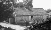 Skräddare Anders Pettersson med kubb och käpp framför ett (sitt?) hus i Grevalid, Grimmared. Stugan är omålad, har sticktak (pärt) och dörren är tvåbladig med fiskbenspanel. (Negativet är skadat)