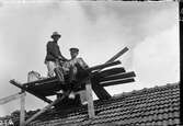 Ett par män murar upp en skorsten på ett tak belagt med flacktegel. På den provisoriska ställningen står hinkar med murbruk och den stående mannen har en murslev i handen.
