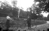 En flicka och en pojke står med varsin cykel på en väg i trädgårdsmiljö. I slänten bakom dem står en flaggstång vid en blomsterrabatt med en kastanj. Längre ner finns en vattenpump. Flickan bär keps och pojken en skärmmössa, samt ett fickur med kedja i västen.