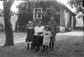 En mor sitter omgiven av fyra barn i trädgården vid ett gårdshus. De två äldsta pojkarna är klädda likadant, medan den minste är klädd i kortbyxor och kolt med sjömanskrage av samma randiga tyg. Flickan har en mörk klänning med vita spetsar på krage, ärmar och nedtill. 
(Se även bild nr GEA134)