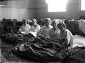 Interiör med fem kvinnor som ligger och vilar under filtar på ett halmtäckt golv.