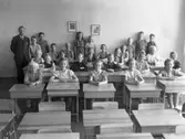Klass 3F i klassrummet på Hagaskolan med lärare Evert Malmqvist till vänster. Några av barnen står vid väggen, övriga sitter i skolbänkarna. 
Längst till höger står Curt-Arne Lernsten och flickan i rutig klänning intill honom heter Gun Johansson. Flickan som sitter två bänkar fram åt vänster från Gun heter Ingrid Linnér. Vid tavlorna står Berit Strannegård Nyberg till vänster och Gun-Britt Bengtson till höger. Främre raden från vänster: Anna-Lisa Karlsson, Elvy, Britt Svensson, ?, Siv Andreasson. Rosmarie Olsson sitter näst längst bak till höger, i ljus klänning. Leif Persson sitter i bakre raden som nr 2 från vänster (barnbarn till Oscar Persson, 