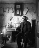 En man sitter vid ett skrivbord där det står en Rikstelefon. Ovanför den hänger en telefonkatalog och buntar med olika dokument. Almanackan med reklam för Pellerins margarin har inte många blad kvar, så troligen är det fredag den 5:e december och då kan året vara antingen 1913 eller 1919.