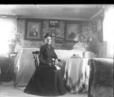 Frederique (Fredrika) Engström sitter med en bok i knät i ett av bostadsrummen i Sällstorps första folkskola. Hon är moster till fotografen, Gunnar Engström, och äldst av de tre systrarna. Porträtten på väggen föreställer förmodligen föräldrarna Carl och Anna Britta Engström.