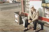 Knut Sund (1914-1998) sitter på en liten fällstol utanför Pressbyrån/dåvarande spelbutiken i Kållereds centrum cirka 1980-tal. I bakgrunden till vänster ses en vit busskur med blå/gul/röda ränder.
