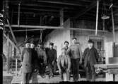 Interiör av fabrik med fabriksarbetare i olika åldrar. I bakgrunden syns remdrift till någon maskin. Två av männen bär kubb, övriga skärmmössor och klädseln varierar.
