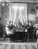 Lärarinnan Berta Engström (fotografens mor) i Sällstorp sitter med fyra skolbarn i sitt eget finrum vid ett bord med duk på. 