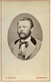 Okänd mans porträtt i oval form, fotograferad i Varberg. Han har skägg och mustasch, ljus väst och kavaj som har en mörk krage.