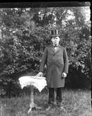 En man iklädd rock och cylinderhatt står i en trädgård vid ett litet pelarbord med duk och håller handen på en uppslagen bok, som ligger på bordet.